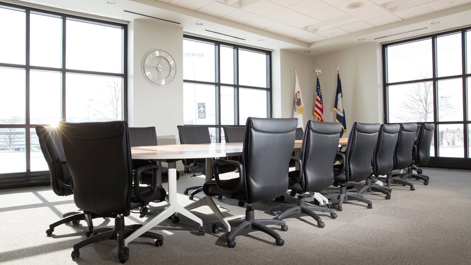 A modern conference room with a large oval table, black chairs, large windows, a wall clock, and two flags.