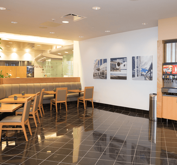 A clean, modern cafeteria with black tiled floors, light gray booth seating, wooden tables and chairs, three framed artworks, and a Coca-Cola soda dispenser.