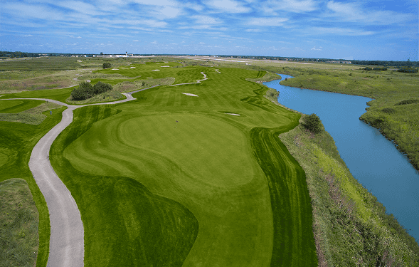 Aerial view of a green golf course with a blue river and an airport in the background under a cloudy sky.