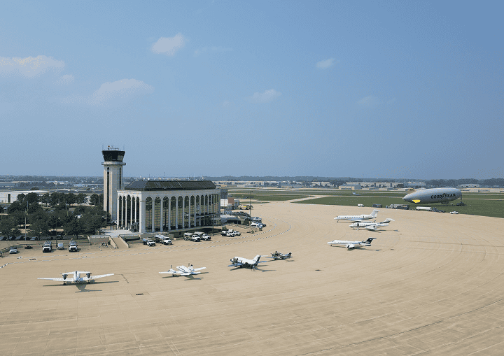 Aerial view of an airport tarmac with a control tower, terminal building, multiple small planes, private jets, and a Goodyear blimp.