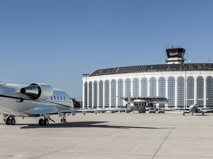 A private jet parked on an airport tarmac in front of a large terminal building with an air traffic control tower under a clear blue sky.