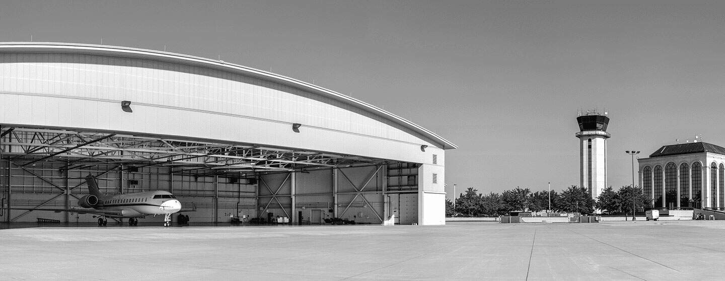 Black and white image of an airport with a private jet in a hangar, a control tower, and a terminal building.
