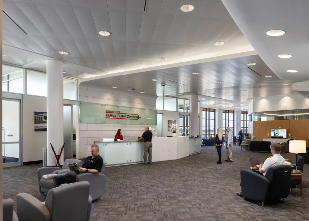 A modern, spacious flight center lobby with a reception desk, multiple seating areas, and several people, including a pilot using a laptop.