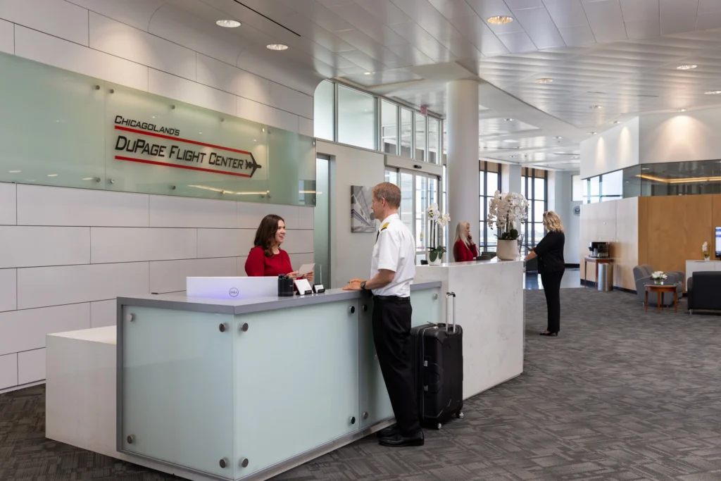 A pilot in uniform stands at a modern reception desk, interacting with a smiling female receptionist, inside the DuPage Flight Center.