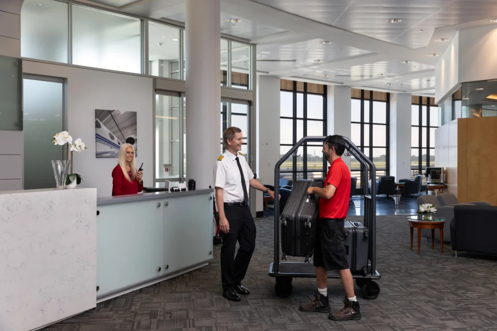 A pilot and baggage handler smiling at each other in a bright airport lounge, with a receptionist behind a desk and a luggage cart nearby.