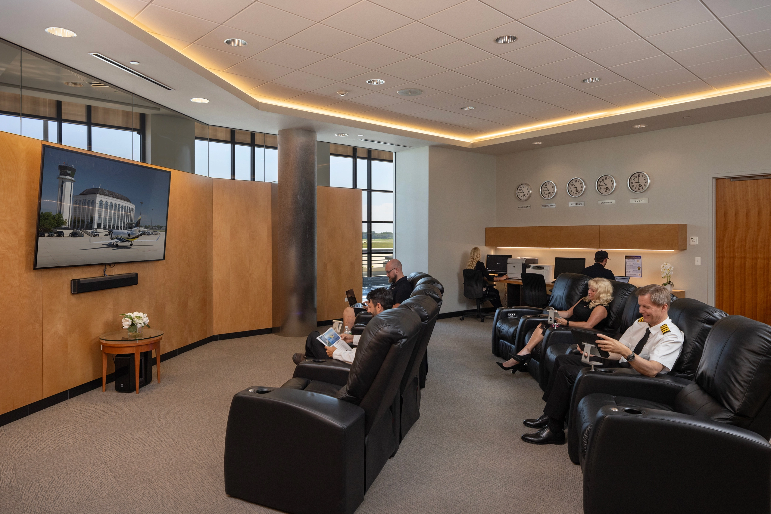 A modern airport lounge with people in black leather recliners, some using tablets or laptops. A large TV shows an airport scene, and wall clocks display different time zones. Large windows overlook an airfield.