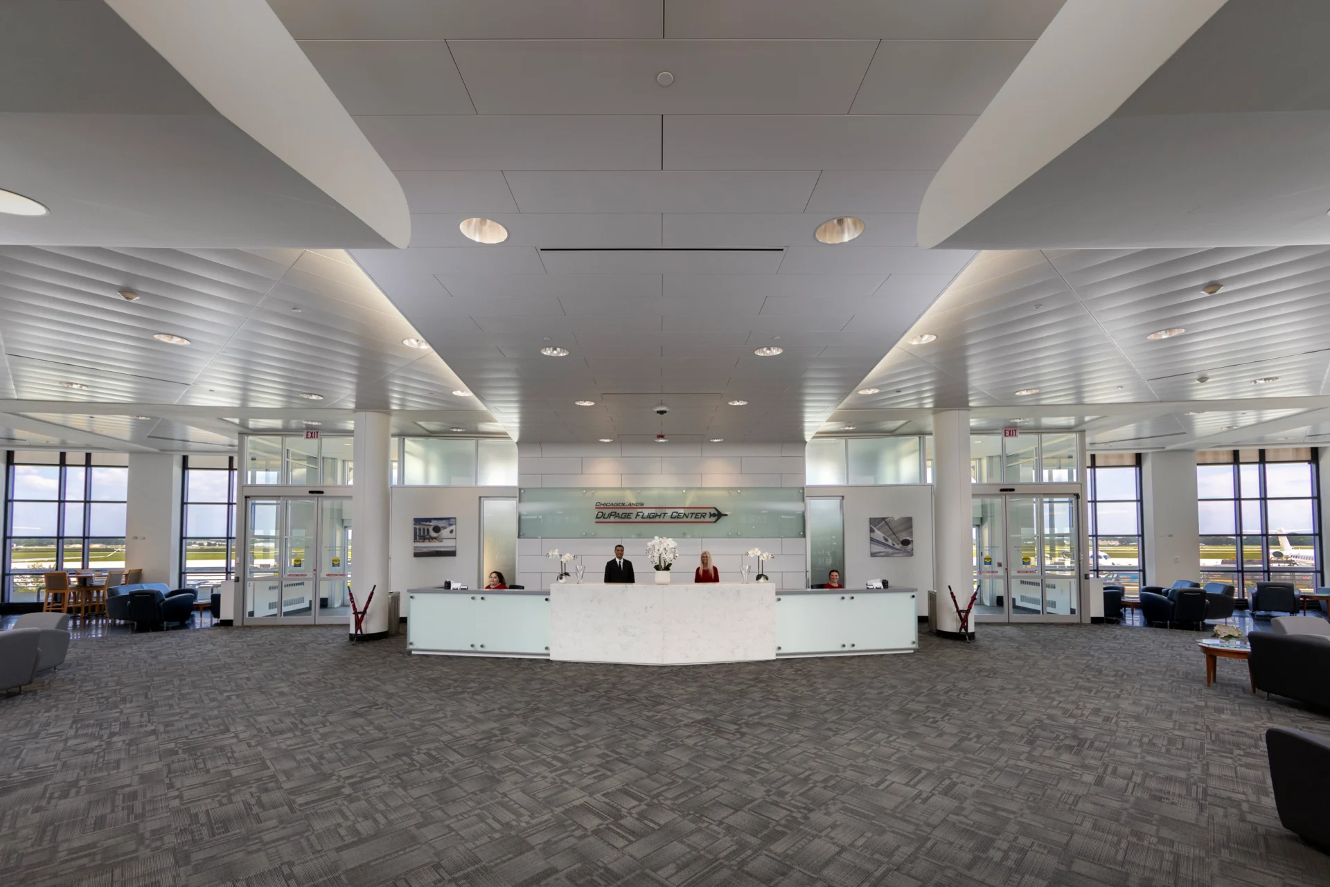 A modern, brightly lit airport reception area with a long, light-colored desk where four staff members are standing. Large windows overlook an airfield with airplanes, and seating areas are visible on both sides.