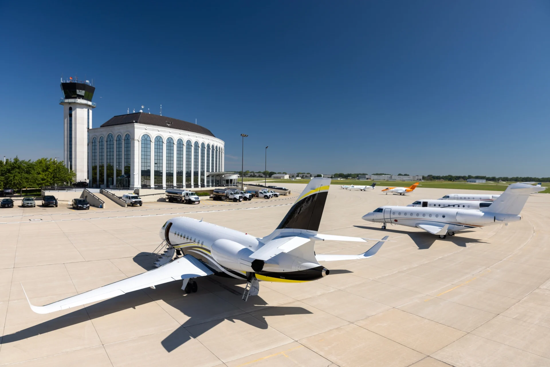 Aerial view of multiple white private jets parked on an airport tarmac next to a white control tower and a large terminal building with arched windows, under a clear blue sky.