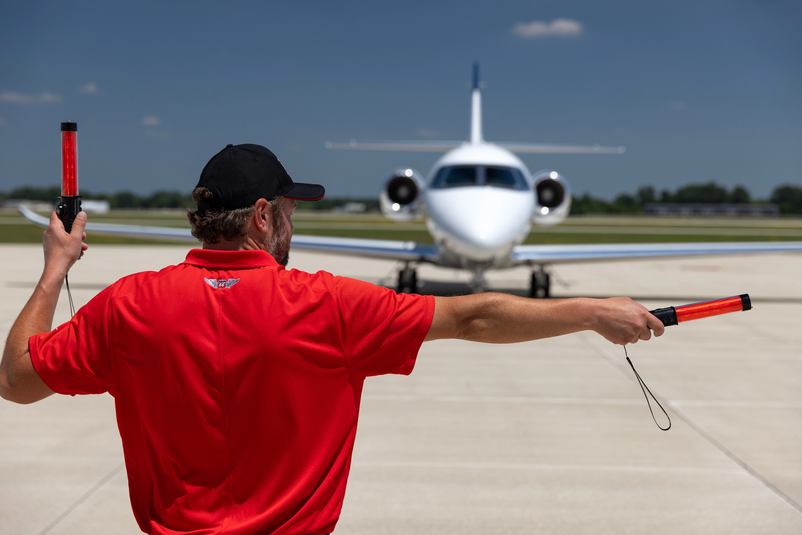 A ground crew member in a red shirt and black cap, seen from behind, directs a white private jet with red marshalling wands on an airfield.