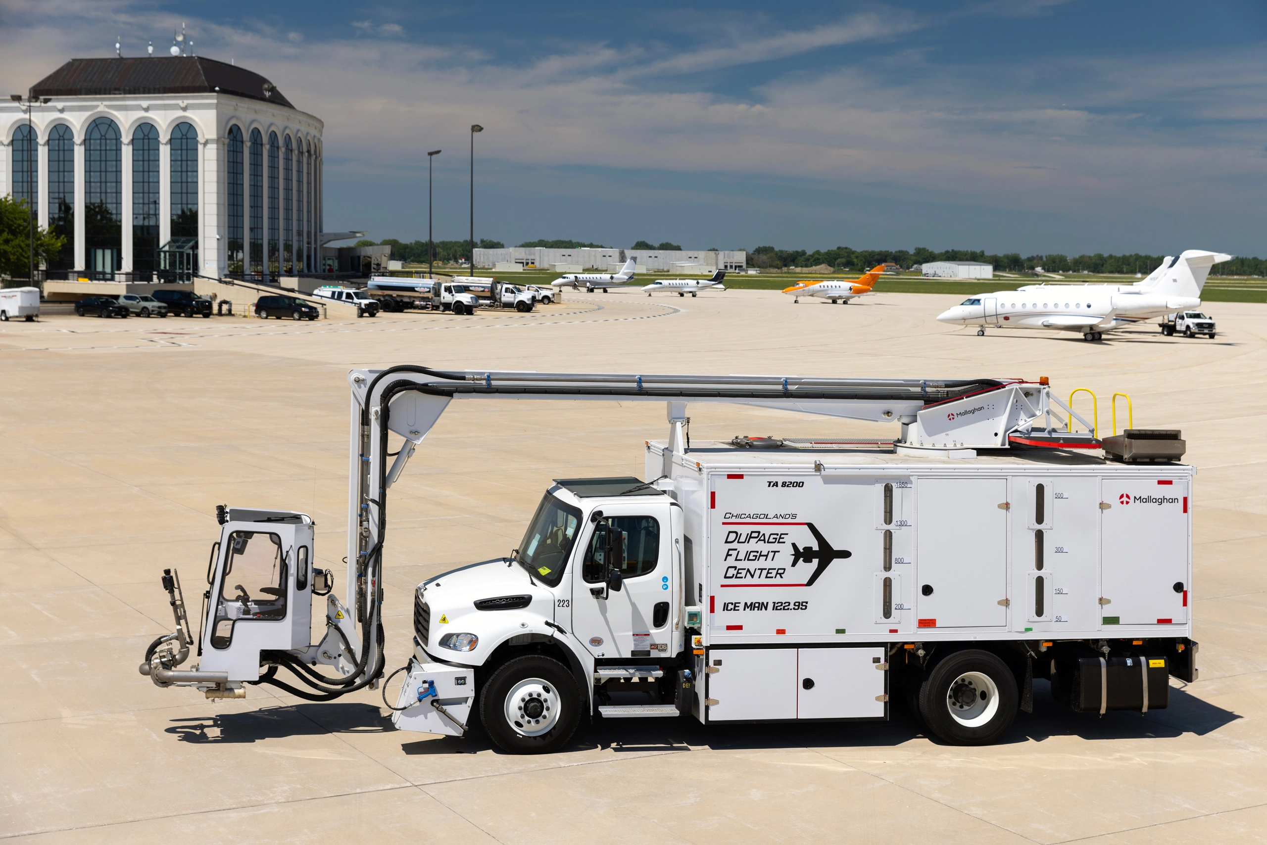 A white Mallaghan de-icing truck with an elevated cabin on an airport apron, with a large building and several private jets in the background.