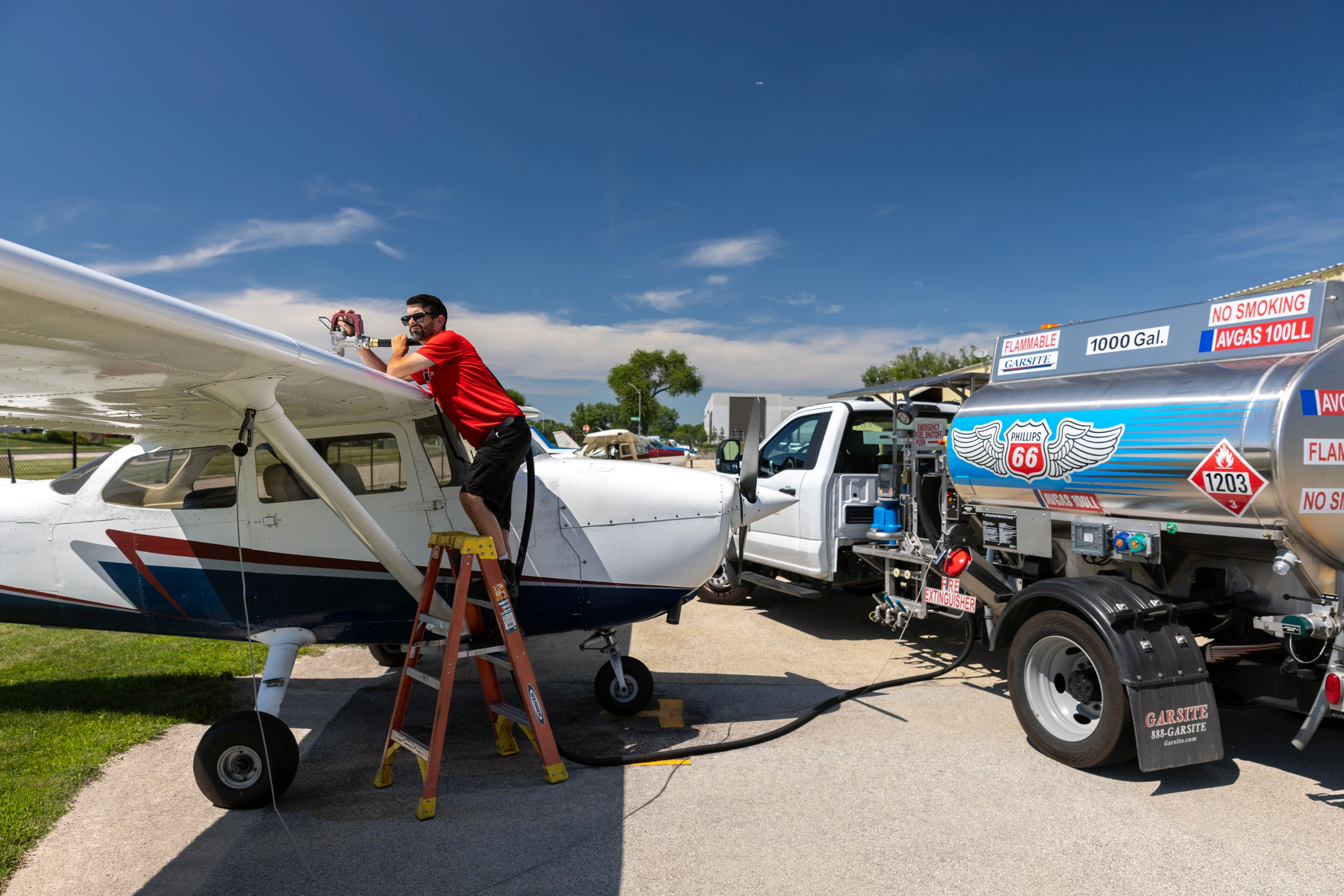 A man on an orange ladder refuels a white airplane from a Phillips 66 fuel truck on a sunny day.