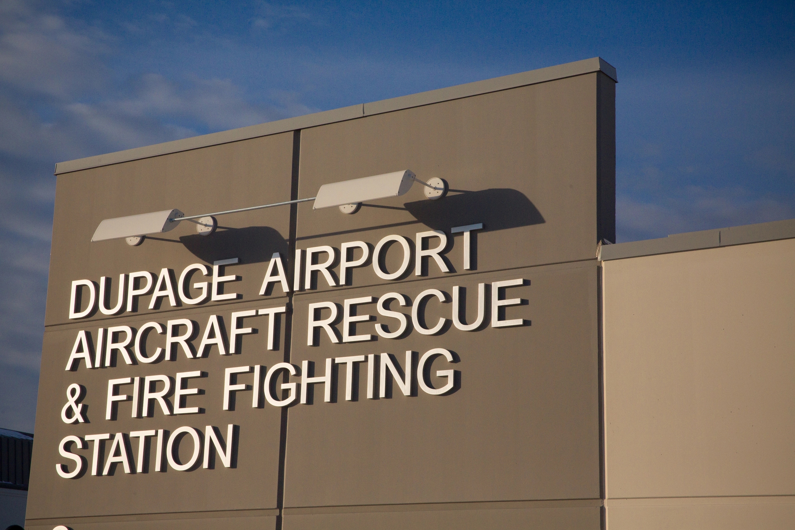 Sign for DuPage Airport Aircraft Rescue & Fire Fighting Station on a tan building under a blue sky.