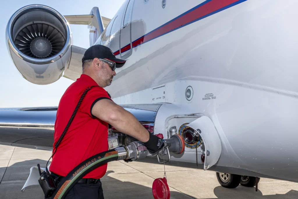 A man in a red shirt and black cap fuels a white private jet with a large hose on an airfield.