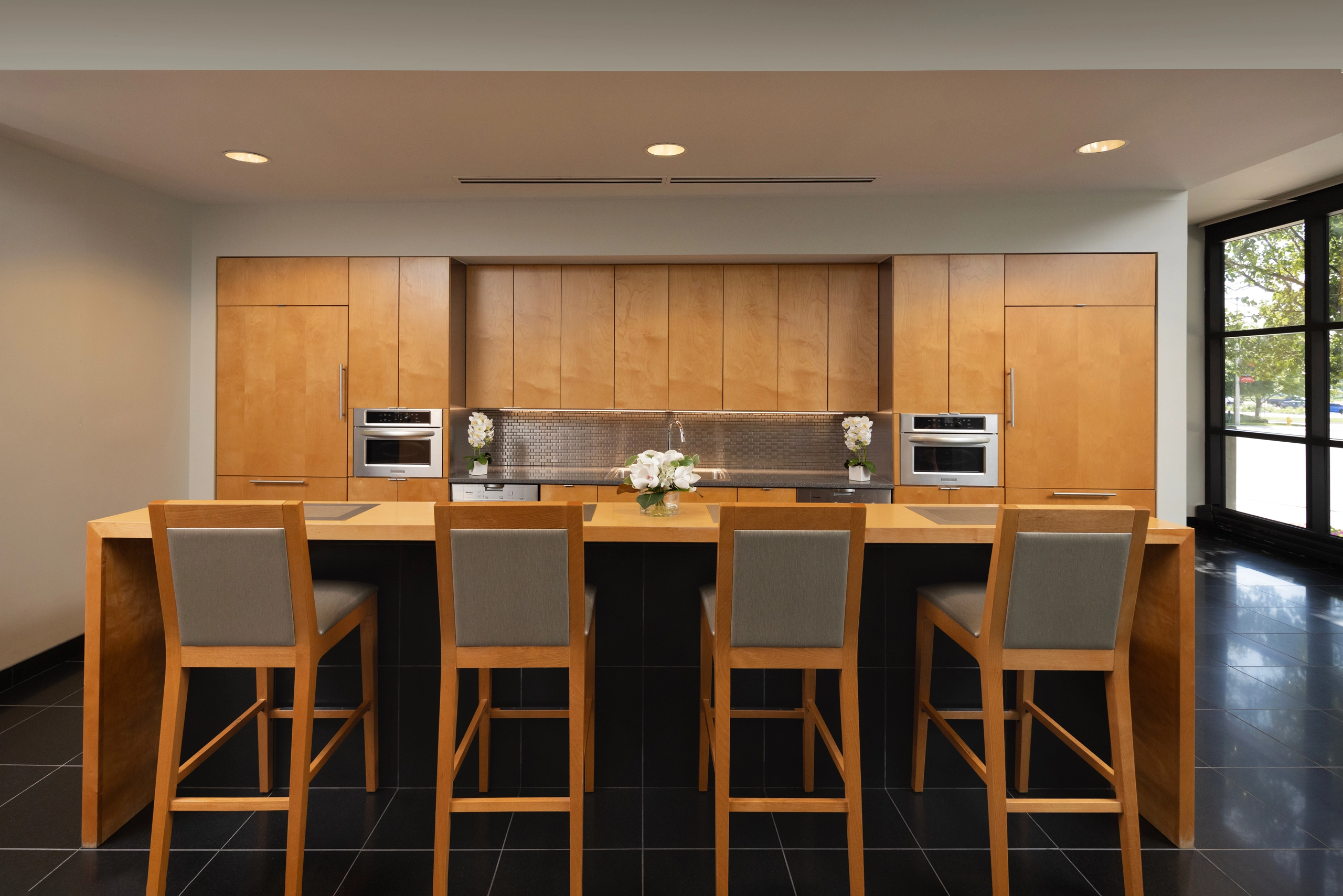 A modern kitchen or breakroom with a light wood island and four gray bar stools. Behind it are light wood cabinets, stainless steel appliances, a tiled backsplash, and a large window.